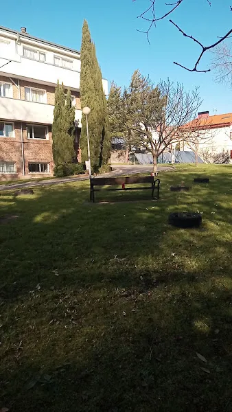 Alai etxe patio ajardinado con banco frente a edificio residencial con césped, árboles y farolas bajo cielo despejado