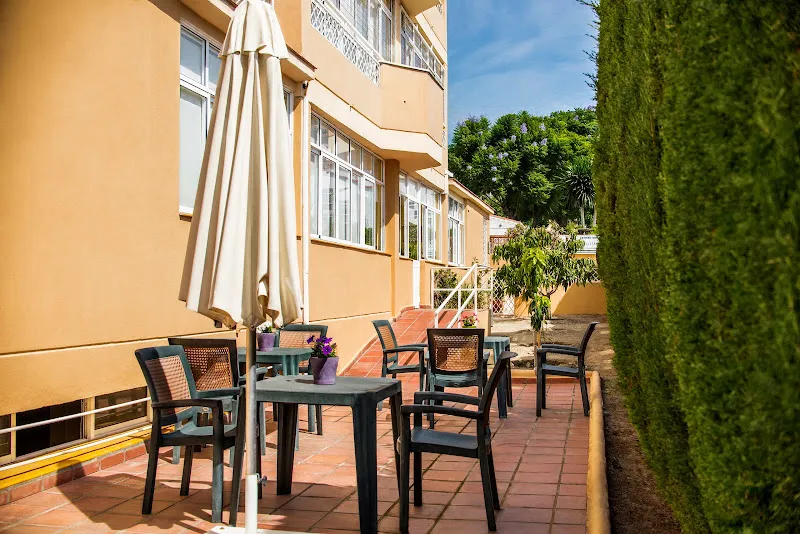 Patio exterior soleado del centro residencial almudena residencia de alzheimer en malaga, espacio de descanso al aire libre con mesas, sillas, sombrilla y macetas junto a fachada y seto