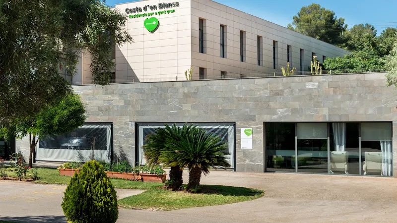 Fachada exterior con grandes ventanales, jardín y cartelería verde frente al edificio domusvi costa d en blanes.