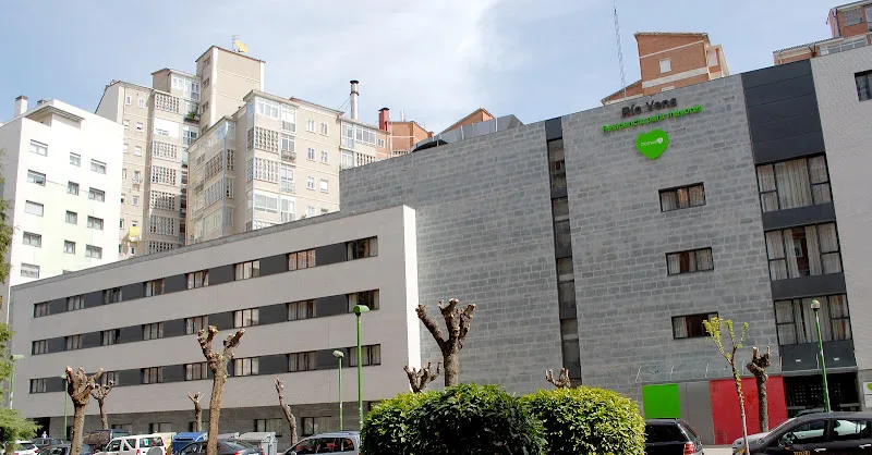 Edificio de fachada moderna con logo verde en forma de corazón de domusvi rio vena junto a árboles podados y coches estacionados