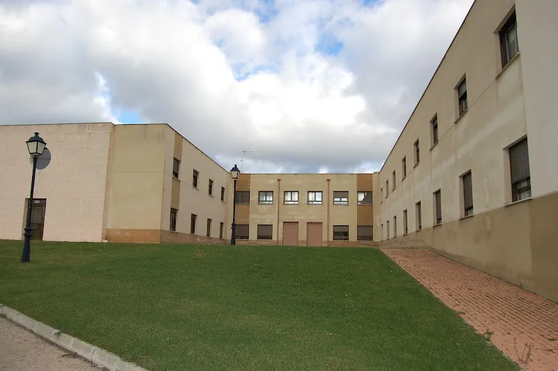 Fachada de la residencia de mayores maria recuenco con césped frontal, farolas y senda peatonal bajo un cielo nublado.