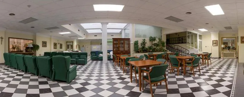 Amplia sala común con sillones verdes, mesas y suelo ajedrezado en residencia geriatrica santa teresa