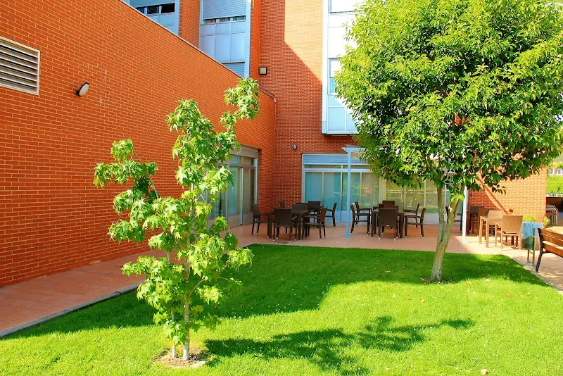 Patio ajardinado con césped, árboles y terraza con mesas y sillas en la residencia para mayores ballesol latina.