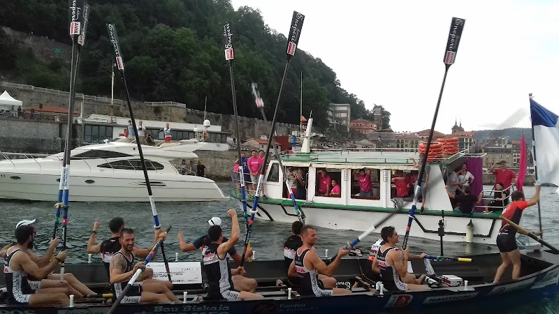 Equipo de remo en el puerto con embarcación de espectadores, imagen ideal para zaindari asistencia a tercera edad